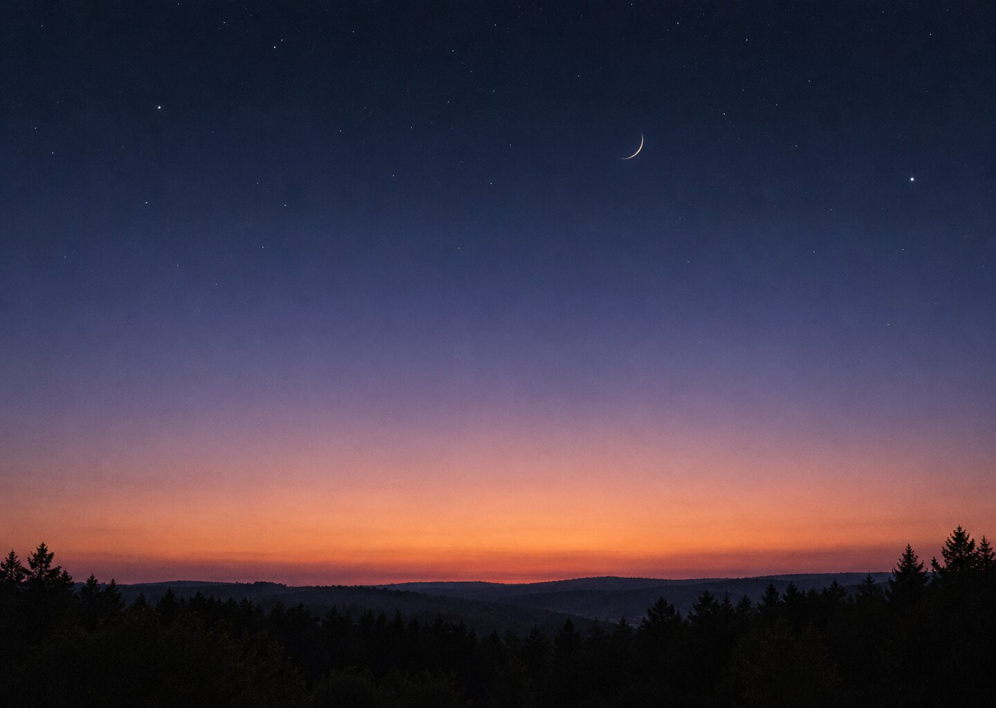A thin crescent moon in a deepening twilight sky above a dark treeline